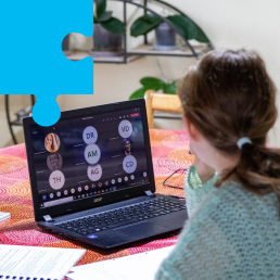 Kobieta pracuje przy domowym stole z kolorowym obrusem. Na stole laptop, na ekranie zdalne spotkanie.