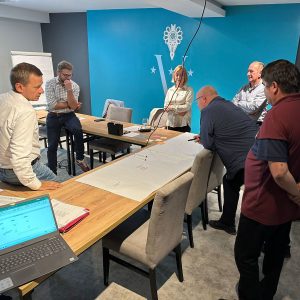 grupa ludzi w sali konferencyjnej stoi nad zapisanymi arkuszami papieru