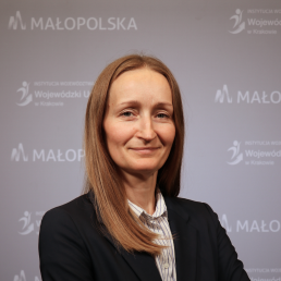 Portret młodej uśmiechniętej kobiety z długimi rozpuszczonymi włosami. Ubrana w elegancką koszulę w paski oraz ciemną marynarkę. W tle ścianka z logo Małopolski oraz Wojewódzkiego Urzędu Pracy w Krakowie.