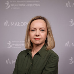 Portret młodej uśmiechniętej kobiety w ciemnej koszuli. Proste jasne włosy sięgają jej do ramion. W tle na ściance logotyp Wojewódzkiego Urzędu Pracy w Krakowie.