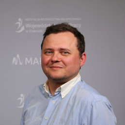 Młody mężczyzna w jasnej eleganckiej koszuli. Ciemne krótkie włosy, delikatny zarost. W tle napisy Małopolska, Wojewódzki Urząd Pracy w Krakowie
