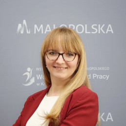 blondynka w średnim wieku, w okularach i bordowej marynarce, uśmiecha się do obiektywu