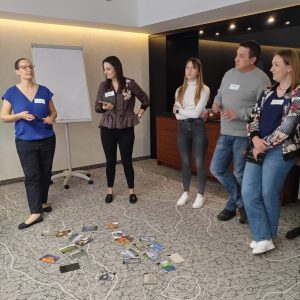 Grupa ludzi stoi w kręgu w sali szkoleniowej. Przed nimi na podłodze leżą rozrzucone karty.