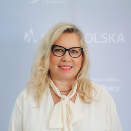 Długowłosa uśmiechnięta blondynka w średnim wieku, w okularach. Ubrana w koszulę z kokardą pod szyją