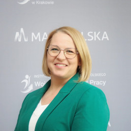 Blondynka, proste włosy do ramion, okulary, jasna bluzka i zielona marynarka