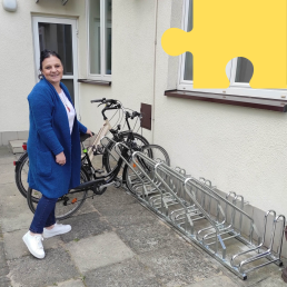 Kobieta w długim niebieskim swetrze i jeansach stoi przy stojaku na rowery, do którego przypięte są dwa pojazdy.