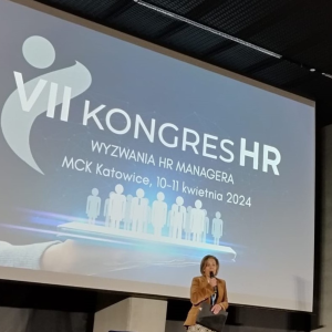 Kobieta na scenie mówi do mikrofonu. W tle na ekranie widoczna nazwa wydarzenia, które prowadzi: VII Kongres HR.