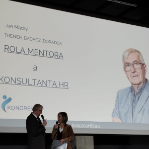 Dwie osoby rozmawiają ze sobą na scenie, stojąc. W tle ekran, na którym jest widoczna sylwetka prelegenta i tytuł jego wystąpienia: Rola mentora a konsultanta HR.