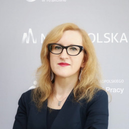 Blondynka w średnim wieku w okularach, długich srebrnych kolczykach, ciemnym topie i żakiecie