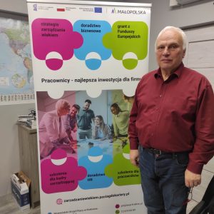 Starszy mężczyzna w bordowej koszuli i jeansach stoi na tle baneru reklamowego projektu "Pracownicy - najlepsza inwestycja dla firmy"