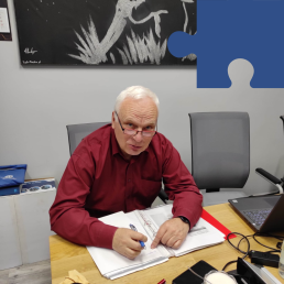 Starszy mężczyzna w bordowej koszuli siedzi przy stole i notuje coś w zeszycie.