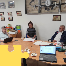 Dwóch starszych mężczyzn i młoda kobieta siedzą przy stole konferencyjnym. Na pierwszym planie widoczny ekran laptopa.