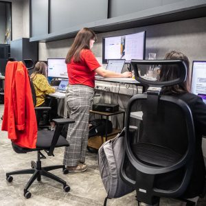 Dwie młode kobiety pracują w open space przed monitorami, jedna na siedząco, druga na stojąco.