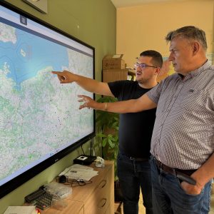 Dwóch mężczyzn rozmawia, stojąc przed dużym ekranem z wyświetloną na nim mapą.