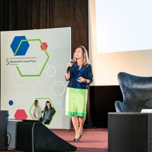 Kobieta w żakiecie i zielonej spódnicy przemawia ze sceny do mikrofonu. W tle baner WUP w Krakowie