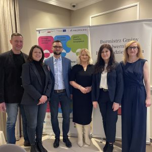 Cztery kobiety i dwóch mężczyzn w eleganckich ubraniach stoi na tle baneru projektu "Pracownicy - najlepsza inwestycja dla firmy" oraz baneru Burmistrza Gminy Alwernia