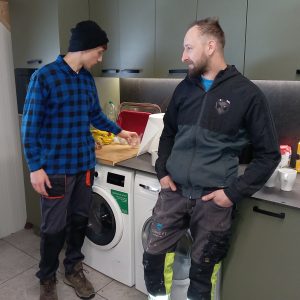 Dwóch młodych mężczyzn w roboczych uraniach stoi w kuchni. Jeden opiera się o blat, na którym leżą akcesoria kuchenne, drugi odstawia szklankę.