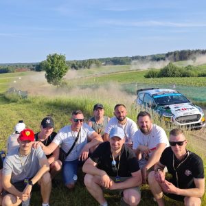 Grupa mężczyzn pozuje do zdjęcia w plenerze, na tle jadącego samochodu rajdowego.