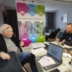 Dwaj mężczyźni w średnim wieku rozmawiają przy stole, na którym widać dokumenty i otwarty laptop. W tle ścianka z napisem: "Pracownicy - najlepsza inwestycja dla firmy".