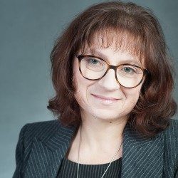 Kobieta w okularach, z grzywką uśmiecha się do obiektywu.