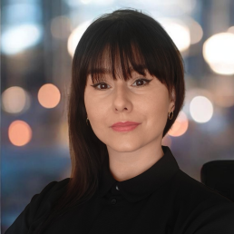 Młoda brunetka z grzywką, w czarnej koszuli z kołnierzykiem, patrzy w obiektyw
