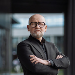 Mężczyzna z brodą, w okularach patrzy w obiektyw. Ma czarną koszulę, brązową marynarkę, stoi z założonymi rękami