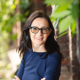 brunetka w okularach i granatowej bluzce uśmiecha się do obiektywu, w tle zielone rośliny