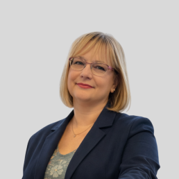 blondynka w okularach z prostymi włosami do ramion. Uśmiecha się do obiektywu, jest ubrana we wzorzystą bluzkę oraz granatową marynarkę