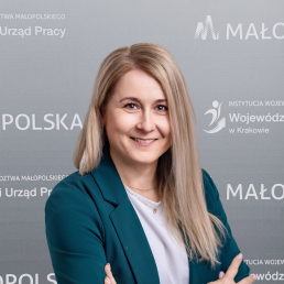 Uśmiechnięta blondynka w białym tshircie i marynarce, na tle ścianki z logotypami Wojewódzkiego Urzędu Pracy w Krakowie oraz Województwa Małopolskiego