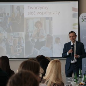 Mężczyzna w granatowym garniturze mówi do mikrofonu na tle prezentacji z napisem: "Tworzymy sieć współpracy".