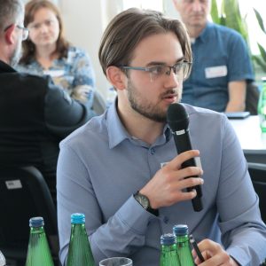 Młody mężczyzna w niebieskiej koszuli i w okularach mówi do mikrofonu.