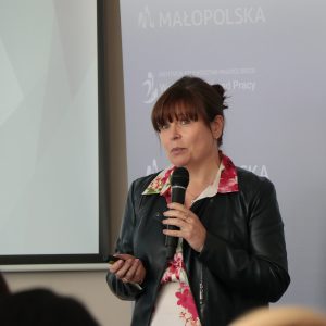 Kobieta w średnim wieku z upiętymi włosami w kolorze kasztanowym mówi do mikrofonu na tle ścianki z logotypem Wojewódzkiego Urzędu Pracy w Krakowie.