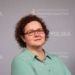 Młoda kobieta w okularach, w jasnej koszuli, z krótkimi kręconymi włosami do ramion. W tle napisy Małopolska, Wojewódzki Urząd Pracy w Krakowie