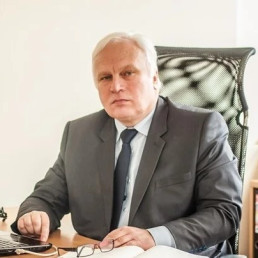 Mężczyzna w garniturze z siwymi włosami siedzi przy biurku, na którym leży laptop, okulary