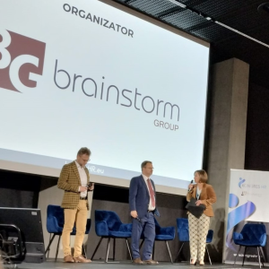 Na scenie stoją 3 osoby. W tle na ekranie widoczny napis: Organizator i logo Brainstorm Group.