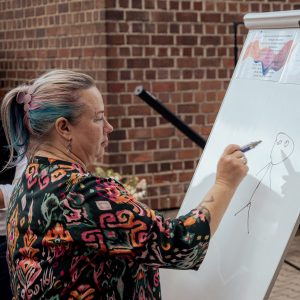 Kobieta w średnim wieku z kolorowymi włosami rysuje na flipcharcie ludzika.