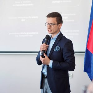 Młody mężczyzna w okularach mówi do zebranych na tle prezentacji.