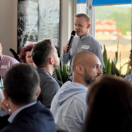 Młody mężczyzna stoi z mikrofonem w ręce wśród uczestników spotkania.