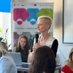 Młoda kobieta w jasnym ubraniu i krótkimi włosami mówi do mikrofonu.