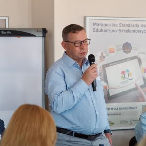 Mężczyzna w średnim wieku w niebieskiej koszuli przedstawia się uczestnikom spotkania.