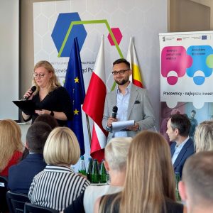 Przed uczestnikami spotkania stoją prowadzący spotkanie: kobieta i mężczyzna. Kobieta mówi do mikrofonu.