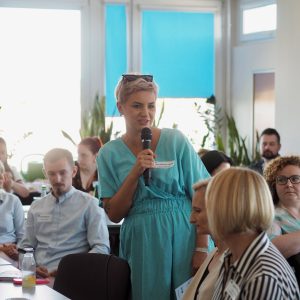 Kobieta w średnim wieku w jasnej niebieskiej sukience mówi do mikrofonu.