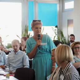 Kobieta w średnim wieku w jasnej niebieskiej sukience mówi do mikrofonu.