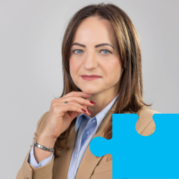 Młoda kobieta z długimi włosami w beżowej marynarce i błękitnej koszuli opiera brodę na dłoni i uśmiecha się do obiektywu.