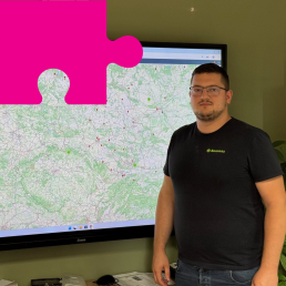 Młody mężczyzna w okularach i czarnym T-shircie z napisem Sowosz stoi na tle dużego ekranu, na którym wyświetlona jest mapa.