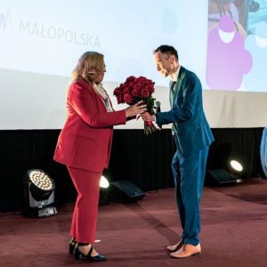 Na scenie kobieta w czerwonym garniturze przyjmuje bukiet czerwonych róż od mężczyzny w granatowym garniturze