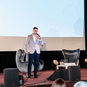 Na scenie mężczyzna w ciemnych spodniach i jasnej marynarce przemawia z mikrofonem w ręce