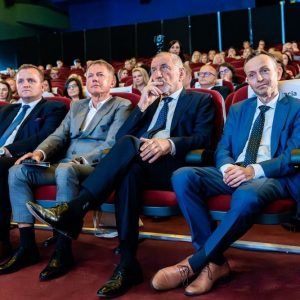 Czterech eleganckich mężczyzn siedzących w pierwszym rzędzie na widowni, patrzą przed siebie