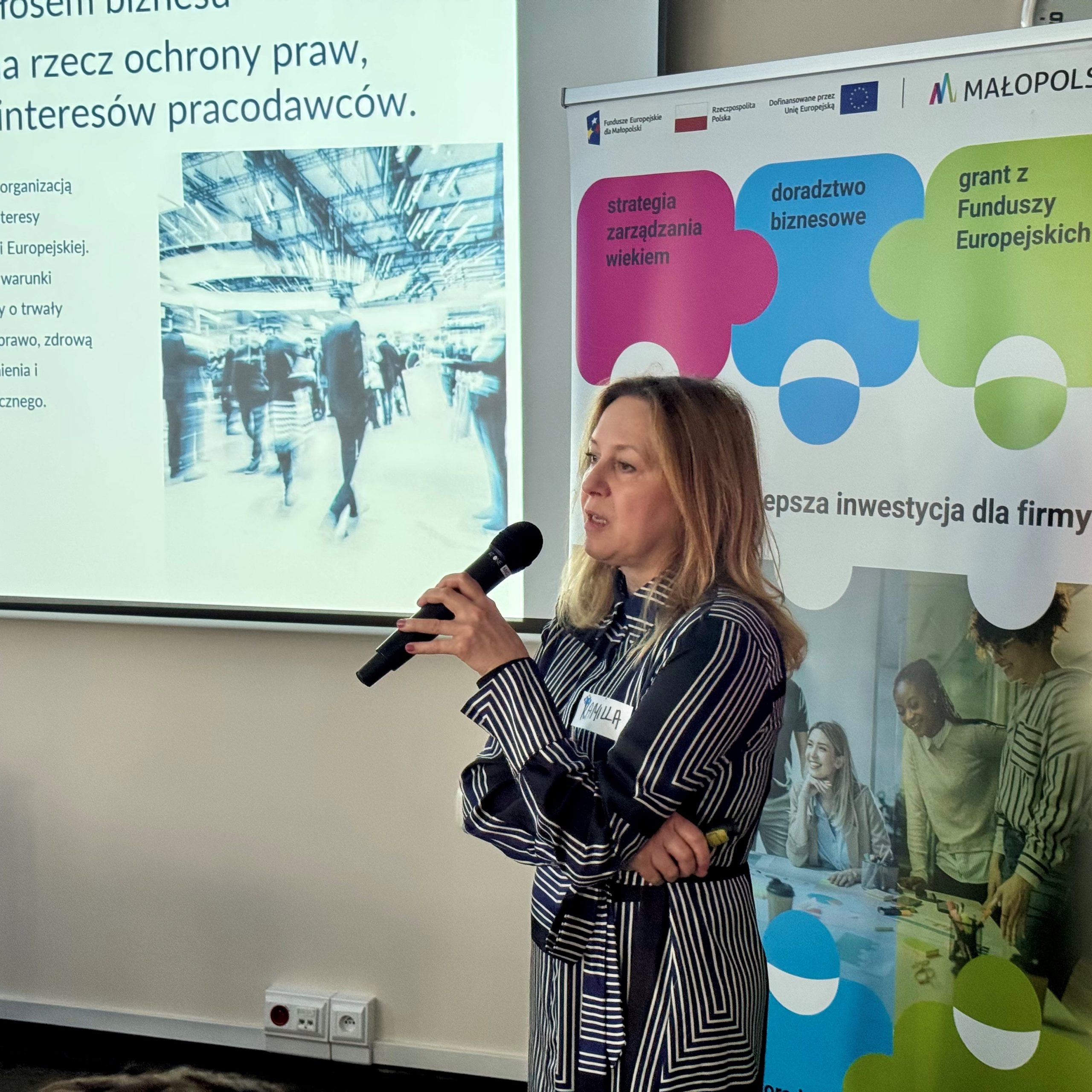 Blondynka w średnim wieku zwraca się do uczestników spotkania. W tle widoczna jest prezentacja.