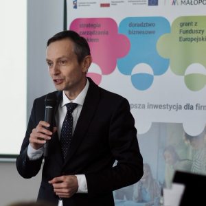 Elegancko ubrany mężczyzna w średnim wieku mówi do mikrofonu na tle baneru projektu "Pracownicy - najlepsza inwestycja dla firmy".